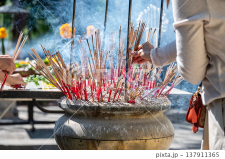 Cropped shot view of woman while place the incense in a pot at Buddhist temple in Thailand. The burned incense may be intended as a symbolic or sacrificial offering to various deities or spirits. 137911365