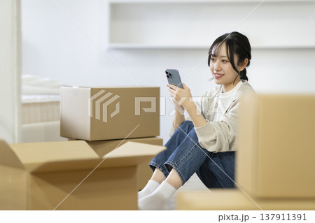 段ボール箱に囲まれて座りスマートフォンを見る若い女性 引っ越し イメージ 段ボール箱に囲まれて座りスマートフォンを見る若い女性 引っ越し イメージ 137911391