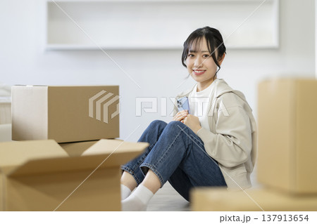 段ボール箱に囲まれて座るスマートフォンを持った若い女性 カメラ目線 引っ越し イメージ 段ボール箱に囲まれて座るスマートフォンを持った若い女性 カメラ目線 引っ越し イメージ 137913654