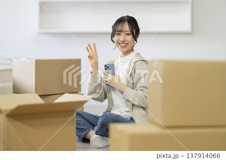 段ボール箱に囲まれて座る笑顔の若い女性　（指3本のポーズ）　カメラ目線　引っ越し　イメージ 137914066