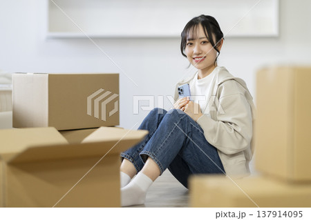 段ボール箱に囲まれて座るスマートフォンを持った若い女性　　カメラ目線　引っ越し　イメージ 137914095