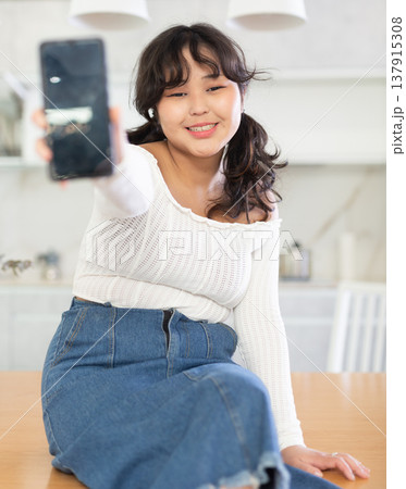 Smiling young girl showing modern smart phone with blank screen, in the living room or in kitchen 137915308