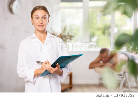 Positive middle-aged masseuse posing with folder in hand in the therapy room 137915358