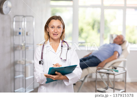 Positive female doctor standing in medical office with document folder Positive female doctor standing in medical office with document folder 137915378