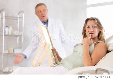 In cosmetology clinic, displeasure client frowns and touches lips, confused doctor in background. 137915454