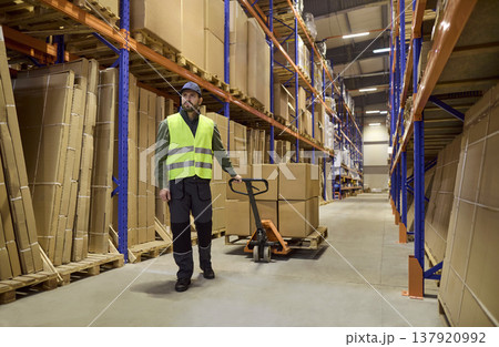 Warehouse worker operating pallet jack moving boxes on industrial storage shelves 137920992