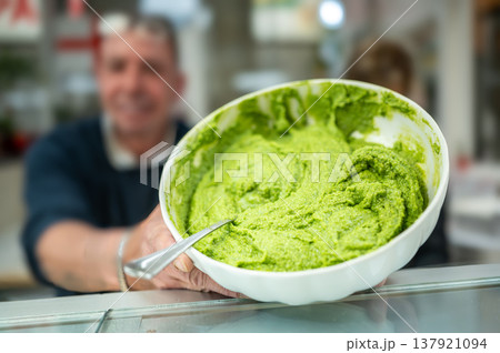 Genoa, liguria, italy. May 18, 2024. Vendor presenting a bowl of vibrant green pesto sauce at mercato orientale, highlighting italian traditional gastronomy 137921094
