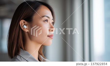 日中の明るい窓際でワイヤレスイヤホンをつけて外の景色を穏やかに眺めている横顔の女性 137922854