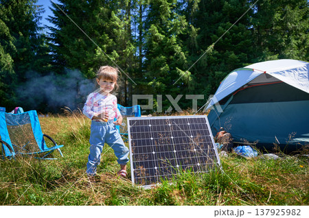 Young child playing near tourist tent with photovoltaic solar panel in summer. On background of dense forest. Integration of renewable energy in outdoor camping activities. 137925982