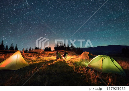 Silhouette of couple hikers resting by campfire under star-studded sky, near illuminated tents at campsite. Night camping in mountains, night sky create magical and peaceful atmosphere. 137925983
