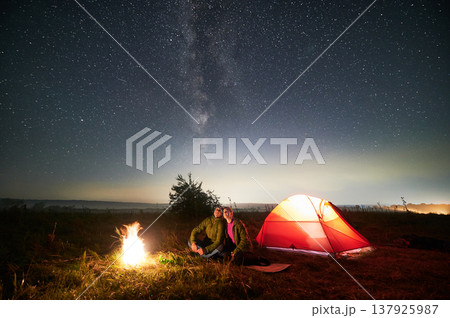 Beautiful view of night starry sky over grassy meadow with couple near campfire. Man and woman hikers sitting near bright bonfire and looking at sky with stars. Concept of hiking and night camping. Beautiful view of night starry sky over grassy meadow with couple near campfire. Man and woman hikers sitting near bright bonfire and looking at sky with stars. Concept of hiking and night camping. 137925987