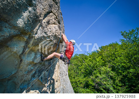 Aerial view of male rock climber ascending rugged limestone cliff with harness and rope for safety. Sportsman climbing on vertical large boulder at Dobvush Rocks in Carpathian mountains, Ukraine. Aerial view of male rock climber ascending rugged limestone cliff with harness and rope for safety. Sportsman climbing on vertical large boulder at Dobvush Rocks in Carpathian mountains, Ukraine. 137925988