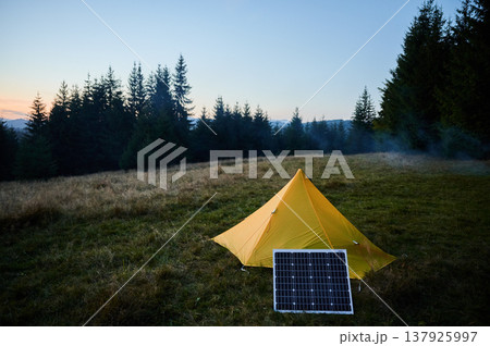 Yellow tent pitched in grassy field with solar panel beside it, capturing last light of setting sun. Backdrop of silhouetted trees and colorful sky emphasizes eco-friendly camping in nature. Yellow tent pitched in grassy field with solar panel beside it, capturing last light of setting sun. Backdrop of silhouetted trees and colorful sky emphasizes eco-friendly camping in nature. 137925997