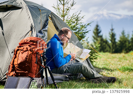 Man sits in tourist tent's entrance, studying map with thoughtful expression. Beside him orange backpack, equipped with tripod and solar panel, set against scenic background of trees and mountains. Man sits in tourist tent's entrance, studying map with thoughtful expression. Beside him orange backpack, equipped with tripod and solar panel, set against scenic background of trees and mountains. 137925998