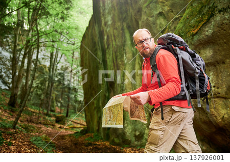 Bearded tourist man studies map while stands in front of large rock in dense forest. Traveler with grey backpack and glasses, planning outdoor adventure, hiking or climbing route. Bearded tourist man studies map while stands in front of large rock in dense forest. Traveler with grey backpack and glasses, planning outdoor adventure, hiking or climbing route. 137926001