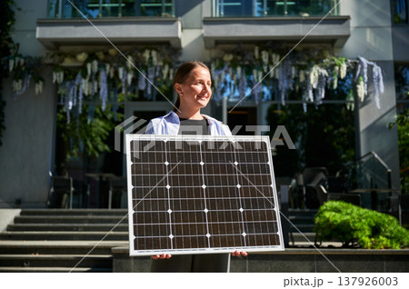 Woman holding solar panel for charging, stands outdoors, smiling in urban setting. Woman holding solar panel for charging, stands outdoors, smiling in urban setting. 137926003