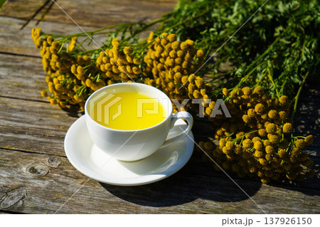 White cup with healthy medicine tansy tea in wooden tray close up. Healthy herbal tansy tea has choleretic and antispasmodic effect. White cup with healthy medicine tansy tea in wooden tray close up. Healthy herbal tansy tea has choleretic and antispasmodic effect. 137926150