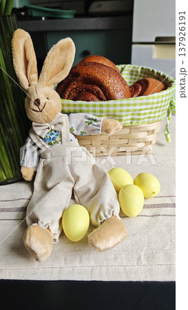 Plush rabbit in overalls is positioned next to yellow eggs and a basket filled with baked goods on a table covered with a striped cloth Plush rabbit in overalls is positioned next to yellow eggs and a basket filled with baked goods on a table covered with a striped cloth 137926191