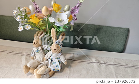 Decorative Easter bunnies in floral attire positioned beside a vibrant bouquet of artificial flowers on a neutral tablecloth in a bright indoor setting 137926372