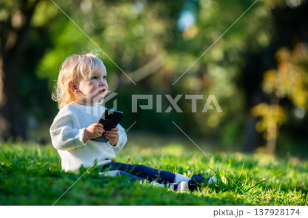 Kid is distracted from his phone while outdoors. A Caucasian child staring away from a smartphone with interest, sitting in a park alone on a sunny day. Cute child sitting in a park on a lovely day. Kid is distracted from his phone while outdoors. A Caucasian child staring away from a smartphone with interest, sitting in a park alone on a sunny day. Cute child sitting in a park on a lovely day. 137928174