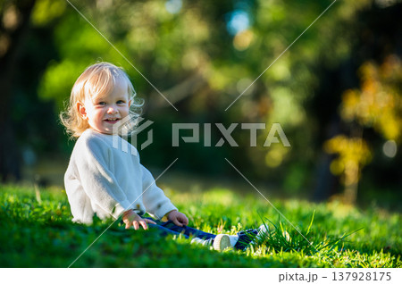 Happy cute toddler enjoying a summers day in a park while sitting on the grass. Smiling adorable boy sitting on the grass in a park on a sunny day. Cheerful small child alone on the lawn and looking Happy cute toddler enjoying a summers day in a park while sitting on the grass. Smiling adorable boy sitting on the grass in a park on a sunny day. Cheerful small child alone on the lawn and looking 137928175