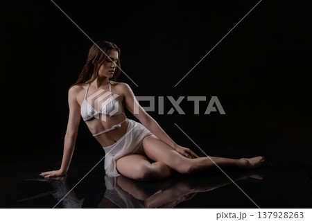 Elegant woman in white swimwear sitting on a wet, reflective surface against a black background. 137928263
