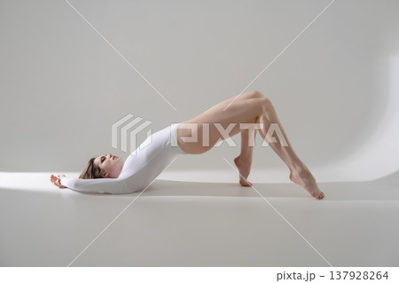 Flexible woman in white bodysuit doing bridge pose exercise on a minimalist gray background. 137928264
