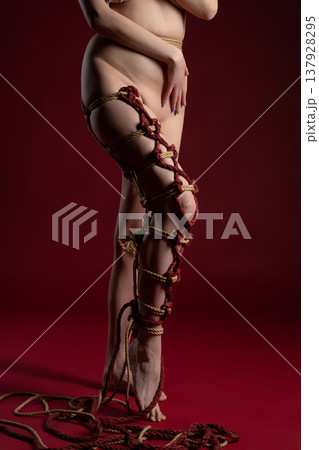 Intricate shibari rope bondage adorns a woman's leg and torso in a dramatic studio setting with a red background. Intricate shibari rope bondage adorns a woman's leg and torso in a dramatic studio setting with a red background. 137928295