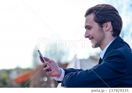 Smiling businessman near a business center with a cell phone. A happy young businessman looking at a smartphone. A young cheerful businessman near a modern tall glass building with a mobile phone. 137929335