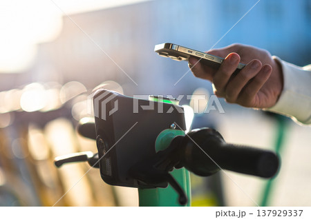Scanning electric scooter QR-code by phone. Pointing a mobile-phone camera to scan a rental scooter's printed code. Using an app to rent an environmentally friendly green electric scooter. Scanning electric scooter QR-code by phone. Pointing a mobile-phone camera to scan a rental scooter's printed code. Using an app to rent an environmentally friendly green electric scooter. 137929337