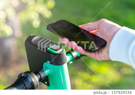 Scanning electric scooter QR-code by phone. Pointing a mobile-phone camera to scan a rental scooter's printed code. Using an app to rent an environmentally friendly green electric scooter. 137929338