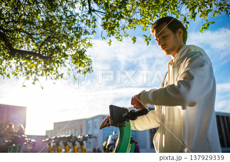 Student looking at his smartwatch while holding onto an electric scooter. Man checks time to make sure that he is not late. Using an app to rent an environmentally friendly green electric scooter. 137929339