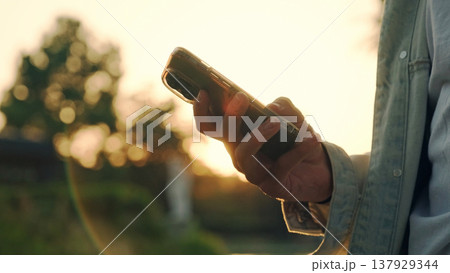 Hands using mobile over sunset. Close-up of men's hands with a mobile in their hands typing sms. Person is typing a text message on a cell phone phone - close-up. People and communication technology. 137929344