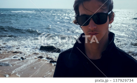 Young man with blown brown hair against the ocean on summer sunny day. Attractive young man looks at the camera on the beach. Handsome guy in black sunglasses poses outdoors on vacation. Young man with blown brown hair against the ocean on summer sunny day. Attractive young man looks at the camera on the beach. Handsome guy in black sunglasses poses outdoors on vacation. 137929734