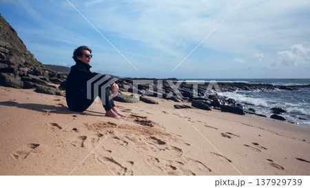 On a sunny summer day, a thoughtful, young guy rests on an empty sandy beach. On a beautiful day, a man sits on a sandy beach. Man in a romantic mood on the beach, looking out at the gorgeous ocean 137929739