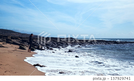 On a hot summer day, a young man wanders barefoot on an empty sandy beach. On a beautiful day, a man walks along a sandy beach. A man in an amorous mood on the beach, admiring nature's beauty.. 137929741