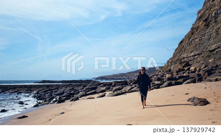 Man with a romantic mood relaxes on the beach contemplating the beautiful nature. Barefoot young man walks on the empty sandy beach on the summer day. Guy is walking on sandy beach on a sunny day. 137929742