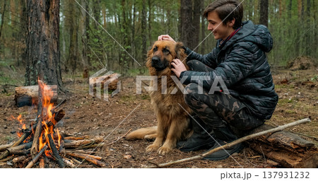 Young man with big dog sits at the fire in the forest. Cheerful guy is stroking his dog, outdoor. Happy caucasian teenager with his German shepherd by the fire in nature, in autumn. 137931232