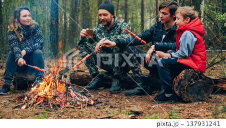Family sits around a campfire on a summer evening. Children with parents are resting in the woods. Family frying sausages over a fire in the woods. Weekend in nature in good company. 137931241
