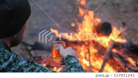 Man with cup of tea the fire in the forest. Human looks at the bonfire and drinks tea on the nature in the summer. Alone traveler sits and rests by the fire with a cup of coffee at dusk. Man with cup of tea the fire in the forest. Human looks at the bonfire and drinks tea on the nature in the summer. Alone traveler sits and rests by the fire with a cup of coffee at dusk. 137931243