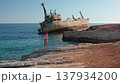 Woman stands on rocky Mediterranean shore near Paphos, Cyprus, gazing at the rusty EDRO III shipwreck grounded off the coast under a clear blue sky, serene travel scene 137934200