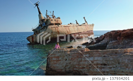 Tourist sitting on a rocky cliff watching the grounded rusty cargo ship EDRO III against the clear blue sky and turquoise Mediterranean Sea near Paphos, Cyprus 137934201