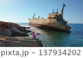 Woman sits on jagged rocks along the Mediterranean coast, gazing at the rusted Edro III shipwreck half-submerged in turquoise water under a clear blue sky in Pegeia, Cyprus 137934202