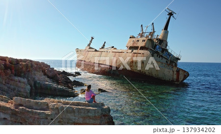 Woman sits on jagged rocks along the Mediterranean coast, gazing at the rusted Edro III shipwreck half-submerged in turquoise water under a clear blue sky in Pegeia, Cyprus Woman sits on jagged rocks along the Mediterranean coast, gazing at the rusted Edro III shipwreck half-submerged in turquoise water under a clear blue sky in Pegeia, Cyprus 137934202