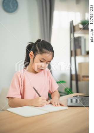 Young Girl Concentrating on Homework in Cozy Indoor Setting, Writing in Notebook While Using Laptop for Study in Bright Room with Natural Light Young Girl Concentrating on Homework in Cozy Indoor Setting, Writing in Notebook While Using Laptop for Study in Bright Room with Natural Light 137934238