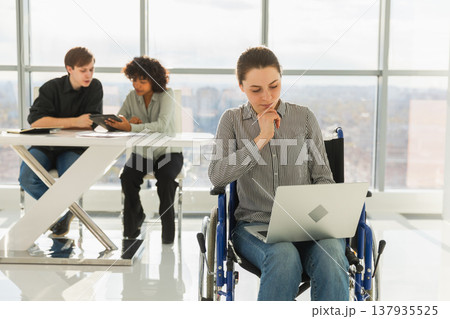 Accessible environment. Woman in chair for people with disability working with laptop in modern office. Diverse multiracial group of people colleagues working together. Young professional team Accessible environment. Woman in chair for people with disability working with laptop in modern office. Diverse multiracial group of people colleagues working together. Young professional team 137935525