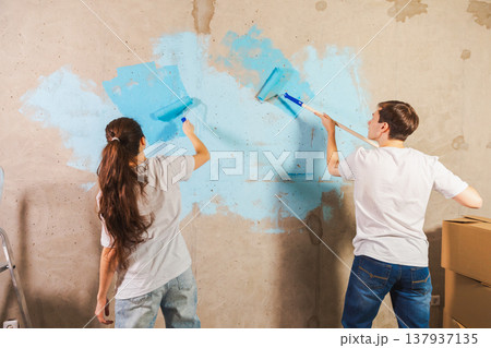 Couple in new home during repair works painting wall together. Happy family holding paint roller painting wall with blue color paint in new house. Home renovation DIY renew home concept Couple in new home during repair works painting wall together. Happy family holding paint roller painting wall with blue color paint in new house. Home renovation DIY renew home concept 137937135