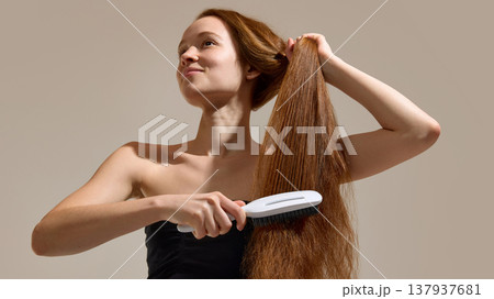 Woman brushing long hair while holding section in studio. Woman brushing long hair while holding section in studio. 137937681