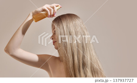 Woman applying spray product to long straight hair in minimal studio environment. Woman applying spray product to long straight hair in minimal studio environment. 137937727