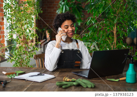 African woman florist gardener working in botanical store using laptop order plants by phone communicating with customer. Female small business owner working at flower shop. Running of own business 137938123